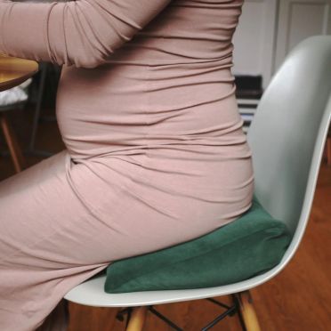 Kobieta w ciąży siedzi na zielonej welurowej poduszce klinowej na białym krześle, drewniana podłoga w tle.